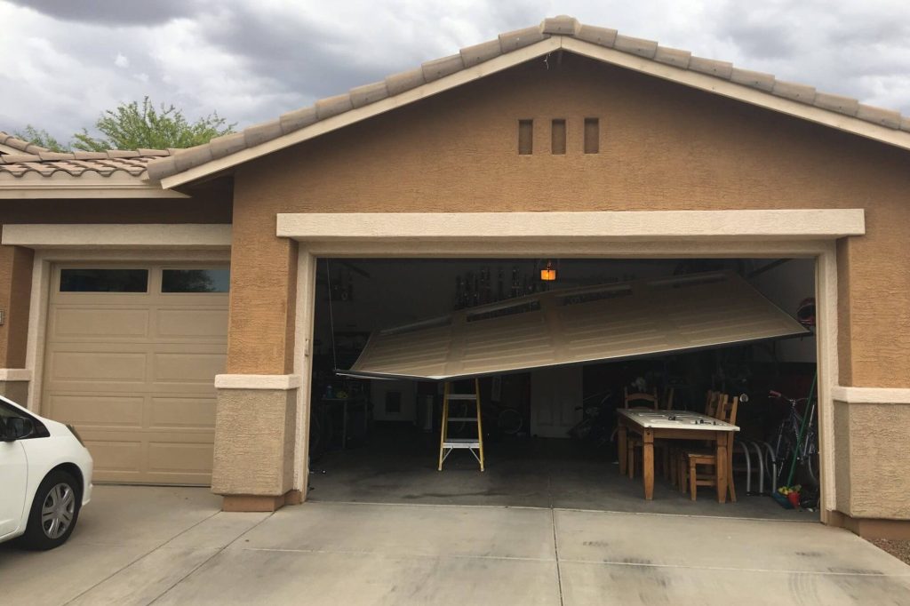 The image shows a garage with a partially broken garage door. The garage door is tilted and appears to be off its tracks, with the left side hanging lower than the right side. Inside the garage, there is a yellow step ladder positioned under the garage door, possibly indicating an attempt to fix it. There is also a wooden table with chairs and various items scattered around the garage. To the left of the broken garage door, there is another closed garage door, and a white car is partially visible in the driveway. The sky above is cloudy, suggesting overcast weather. Associated with Pride Garage Door.
