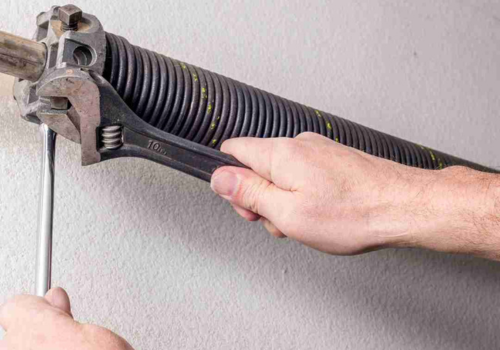 Close-up of a person's hands working on a torsion spring, part of a garage door mechanism. One hand holds a 10-inch adjustable wrench, while the other holds a rod or screwdriver inserted into the winding cone of the spring. The torsion spring is mounted horizontally on a wall or ceiling