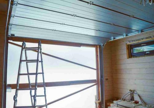 Interior of a garage with a partially open garage door. A metal ladder is positioned near the door, and the garage appears to be under construction or renovation. The garage door is segmented and made of metal panels, connected by hinges and supported by tracks on the ceiling. There is a small window on the right wall, and various construction materials and tools are visible on the floor and shelves. Associated with Pride Garage Door.