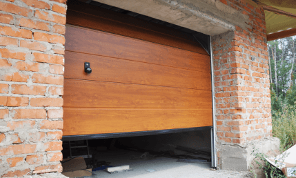 A partially open wooden garage door with horizontal panels installed in a red brick garage. The structure features areas of exposed mortar, while the floor inside the garage is scattered with various items and debris, reflecting a construction or installation process.