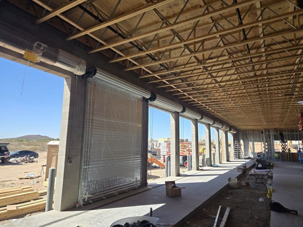 Interior of a construction site with exposed wooden beams, large roll-up doors, and scattered building materials, under a clear blue sky.