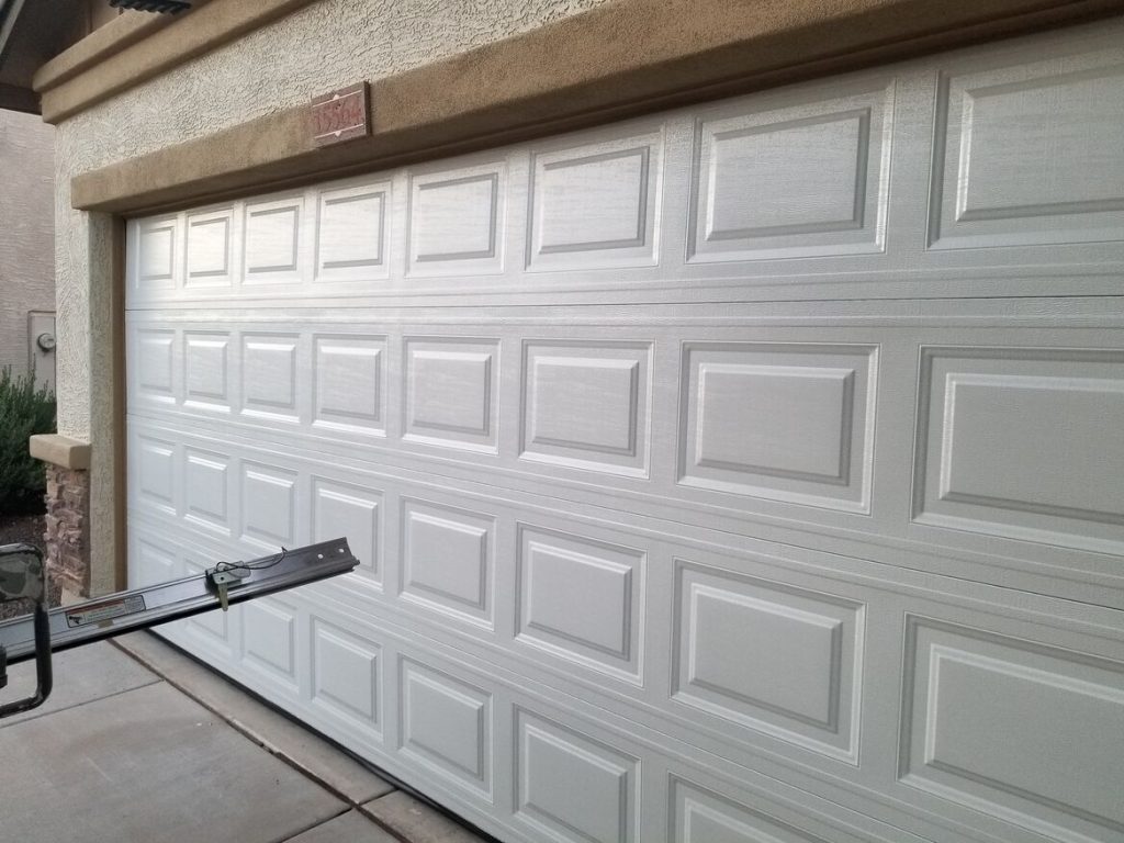 A clean, white garage door with a panel design, shown in a residential setting. A metal tool rests on the driveway.