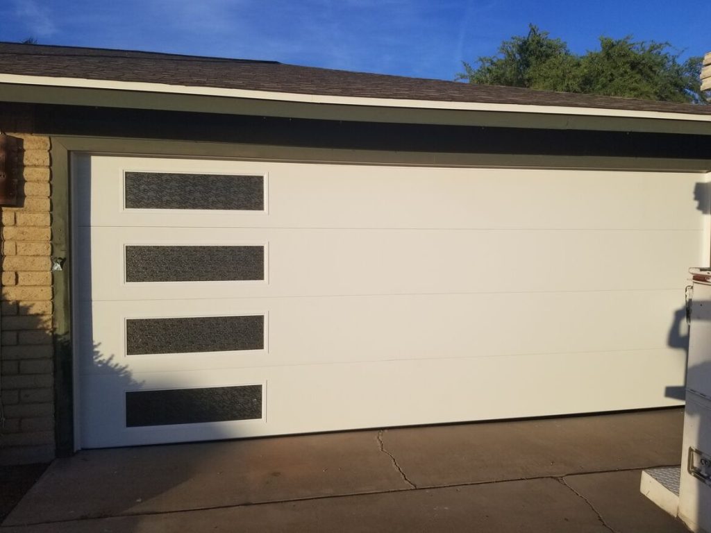 A modern white garage door with three horizontal windows, set against a brick wall and bright blue sky.