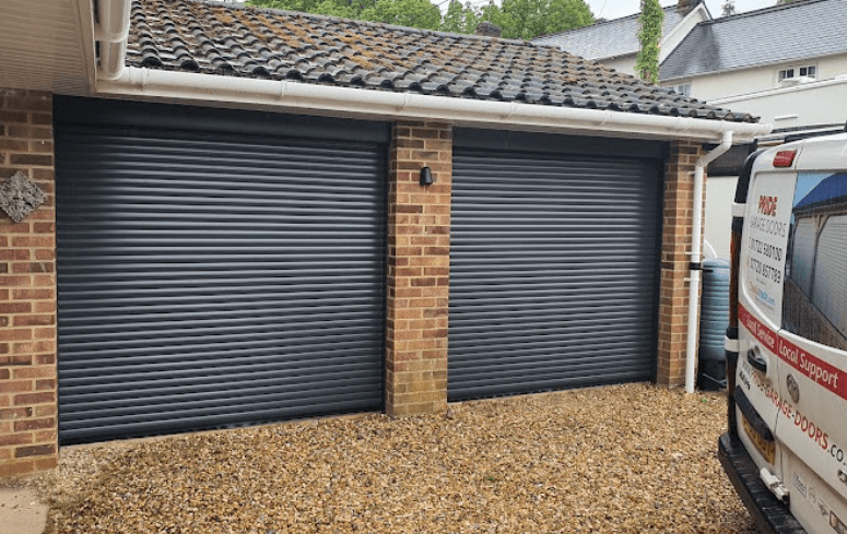 Two black roller garage doors are nestled in a brick wall, beside a van parked on a gravel driveway under a sloped roof.
