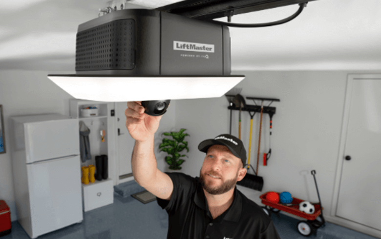 A person adjusts a LiftMaster garage door opener mounted on the ceiling in a tidy garage setting, with various tools and equipment visible.