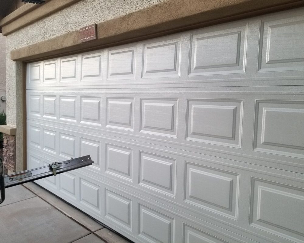 A clean, white garage door with a panel design, shown in a residential setting. A metal tool rests on the driveway.