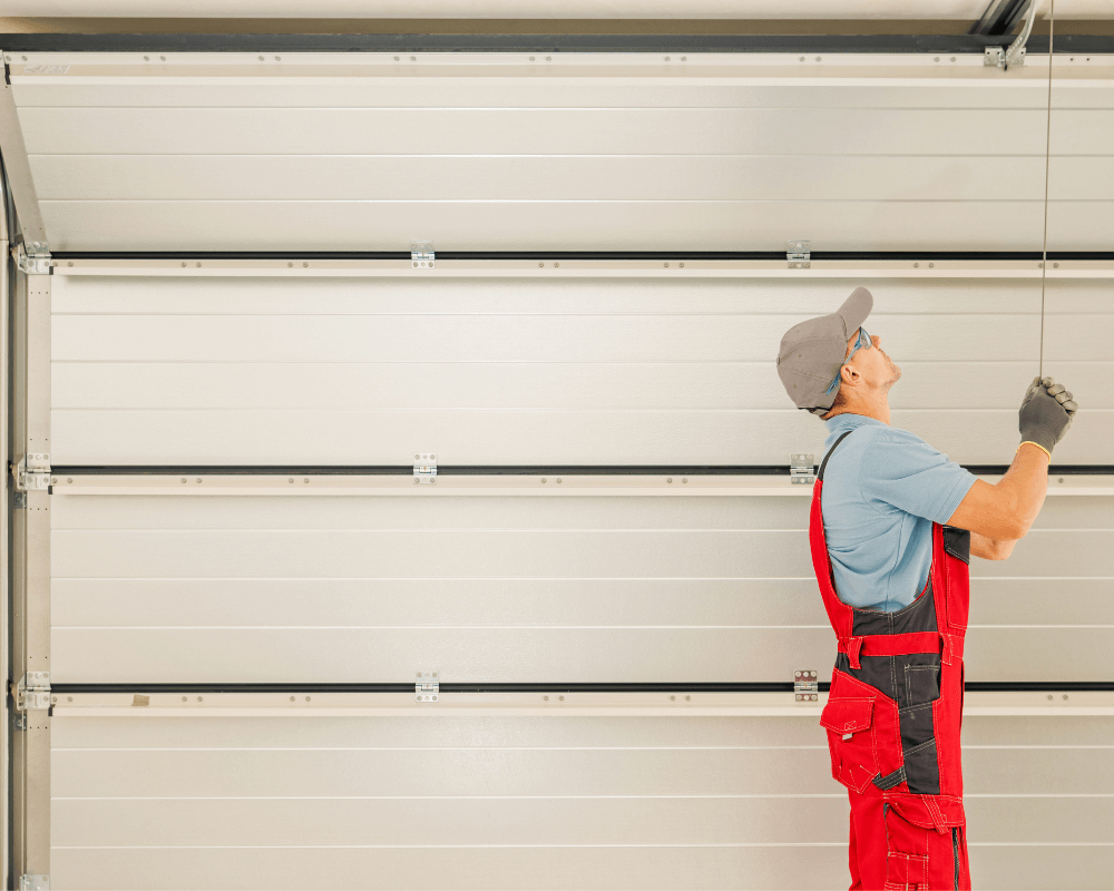 A man in red overalls and gray cap pulls a garage door release cord, with the door visible above him in a brightly lit garage.