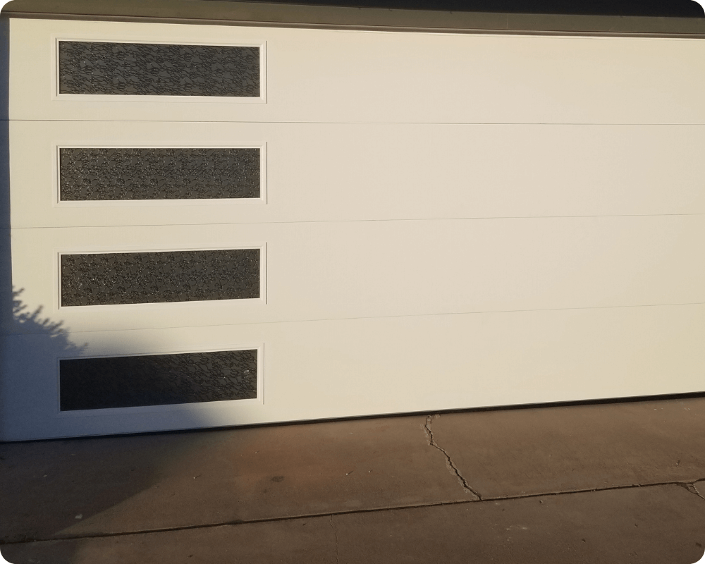 A white garage door with three decorative, dark glass panels at the top and a smooth surface beneath, on a concrete driveway.