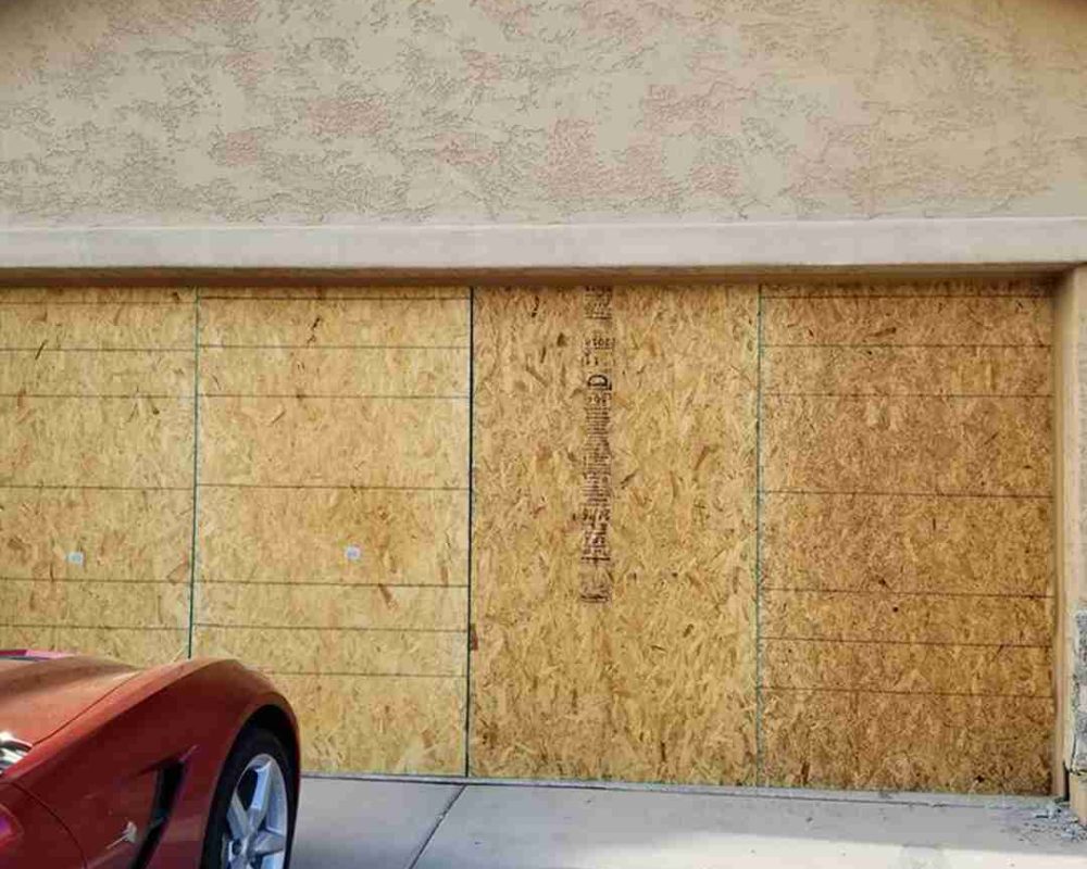 The image shows a garage door that has been boarded up with plywood sheets. The garage door appears to be a double door, and the plywood sheets cover the entire surface. Two circular vents are above the garage door on the wall, and a red car is partially visible in the lower left corner, parked in front of the garage. The plywood sheets have some markings and lines on them, possibly indicating measurements or cuts. Associated with Pride Garage Door.