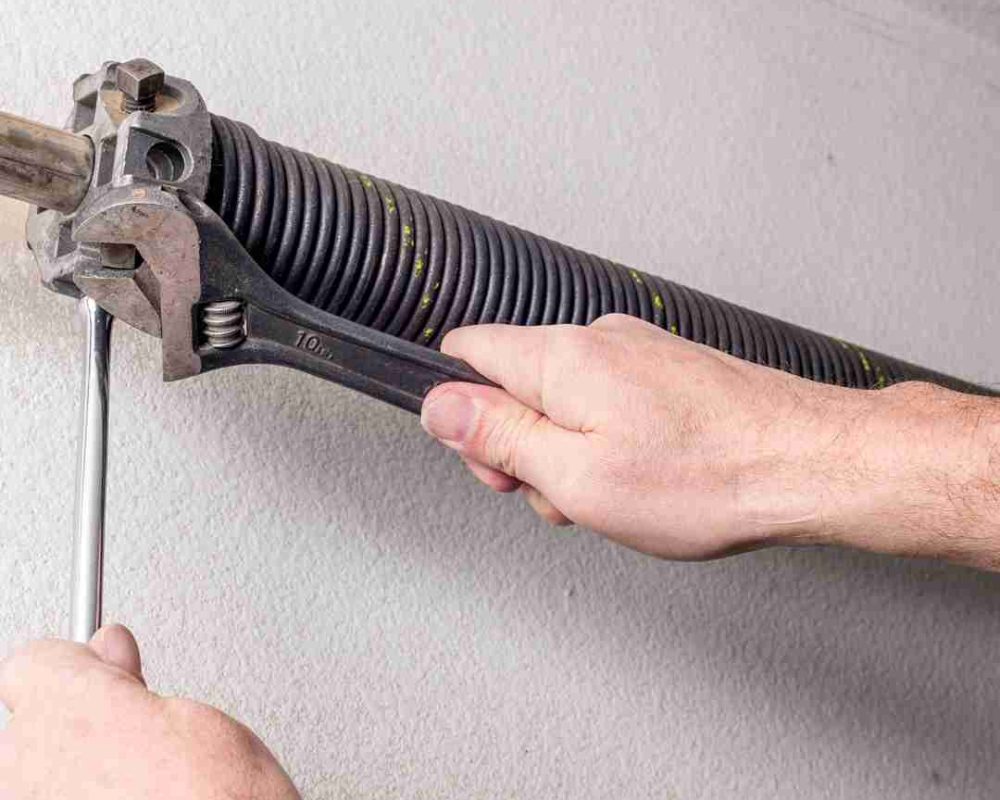 Close-up view of a garage door mechanism, showing a torsion spring mounted on a metal rod, brackets securing the rod to the ceiling, and a black metal bar connected to the garage door opener mechanism. A person is holding a tool and a red and white rope, likely adjusting or repairing the garage door system. Associated with Pride Garage Door.