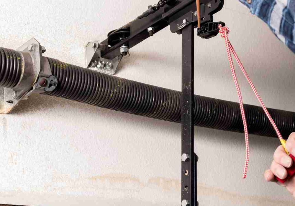 Close-up view of a garage door mechanism, showing a torsion spring mounted on a metal rod, brackets securing the rod to the ceiling, and a black metal bar connected to the garage door opener mechanism. A person is holding a tool and a red and white rope, likely adjusting or repairing the garage door system. Associated with Pride Garage Door.