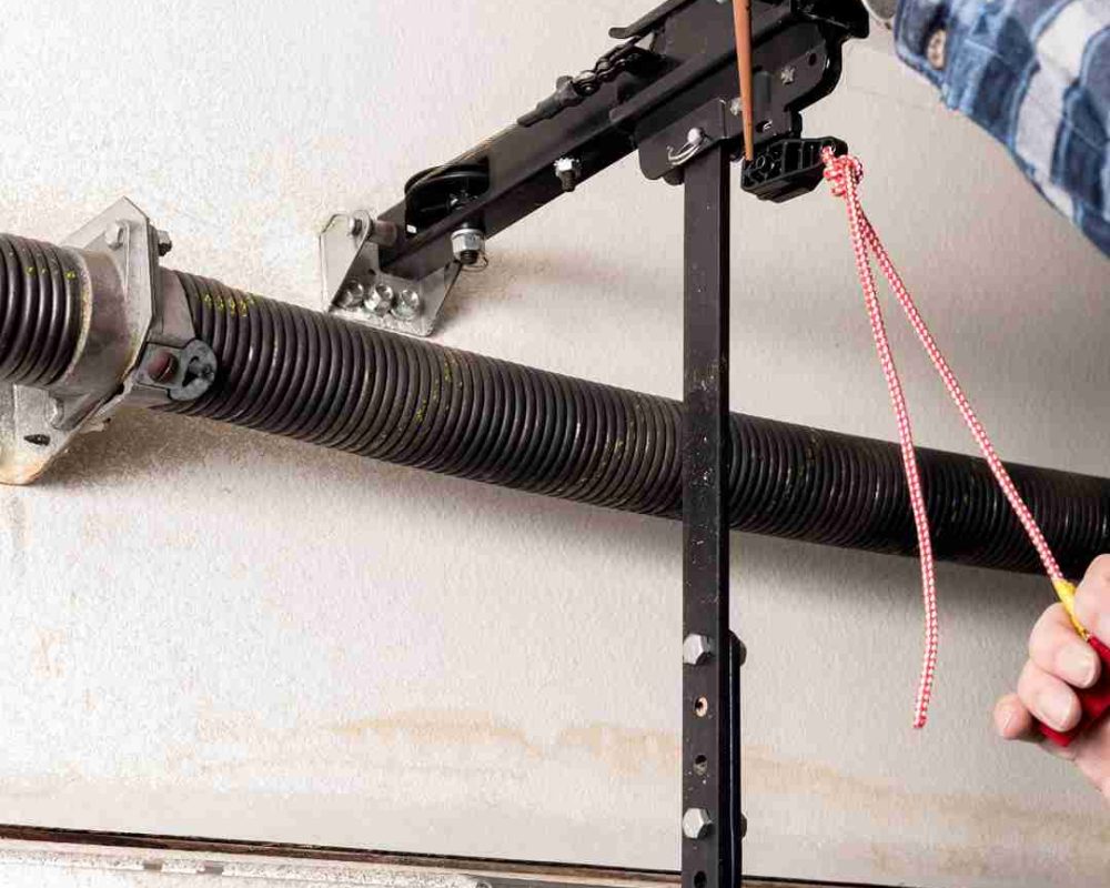 Close-up view of a garage door mechanism, showing a torsion spring mounted on a metal rod, brackets securing the rod to the ceiling, and a black metal bar connected to the garage door opener mechanism. A person is holding a tool and a red and white rope, likely adjusting or repairing the garage door system. Associated with Pride Garage Door.